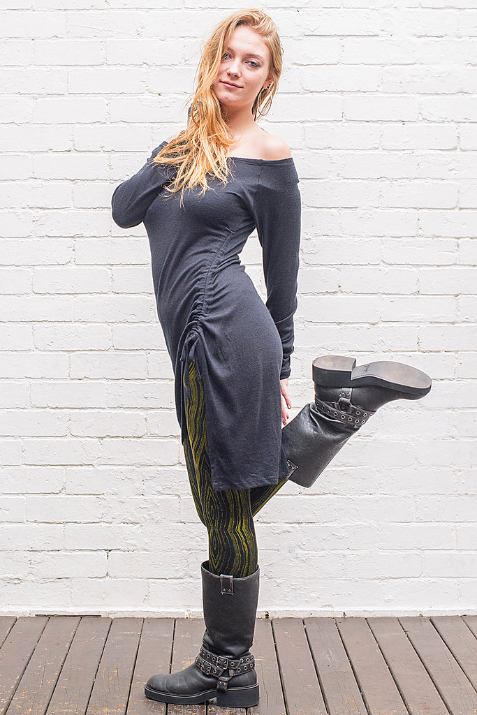 Model wearing a Khione knee length knit dress with boat collar off the shoulder and adjustable assymetric hemline in black from side