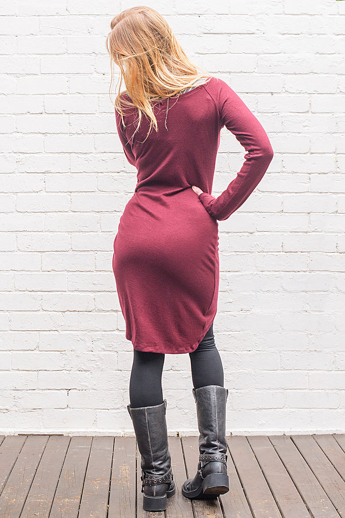 Model wearing a Khione knee length knit dress with boat collar and adjustable asymmetric hemline in maroon from back