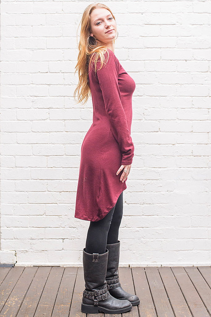 Model wearing a Khione knee length knit dress with boat collar and adjustable asymmetric hemline in maroon from side