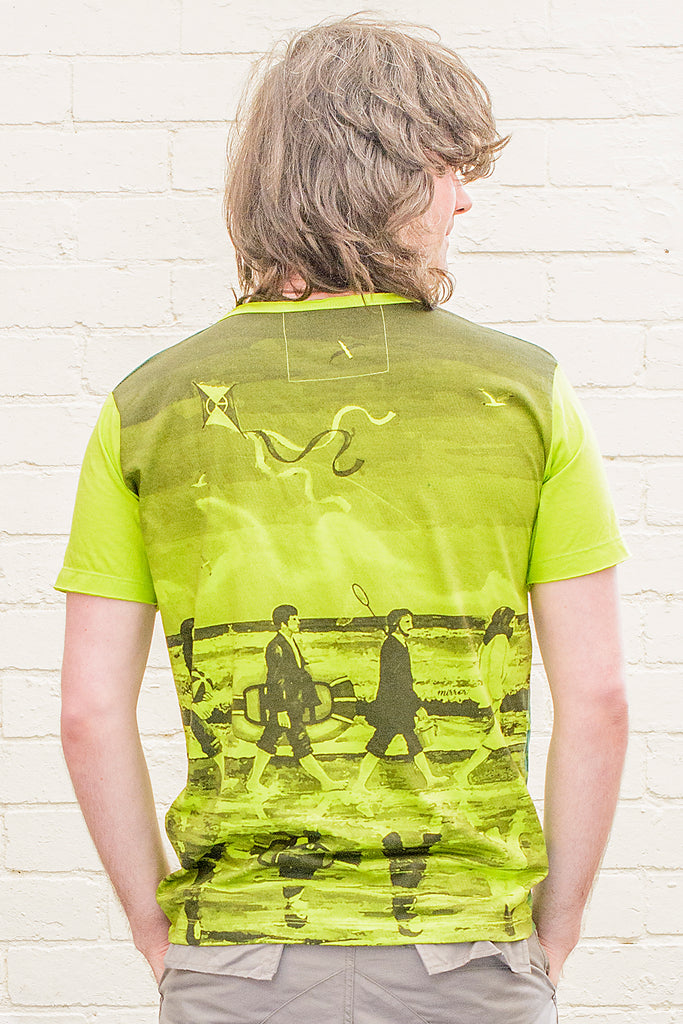 model wearing a bright green T-shirt featuring a painterly design of the beatles walking along a beach with beach toys from the back