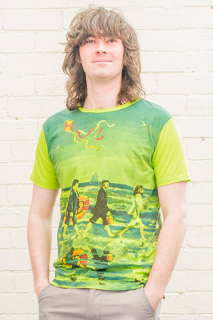 model wearing a bright green T-shirt featuring a painterly design of the beatles walking along a beach with beach toys from the front