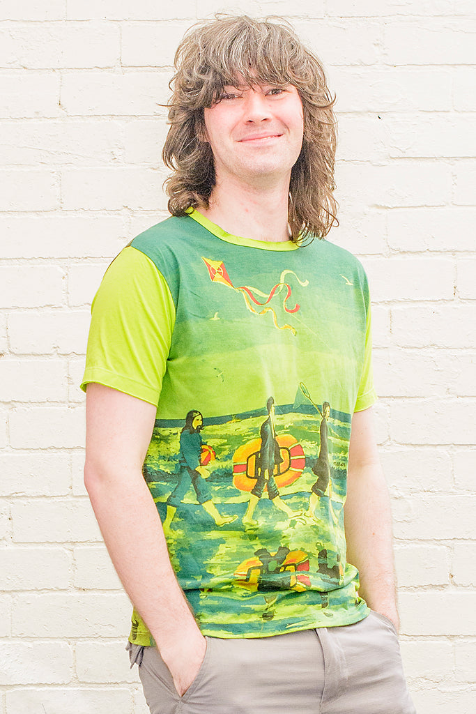 model wearing a bright green T-shirt featuring a painterly design of the beatles walking along a beach with beach toys from the front