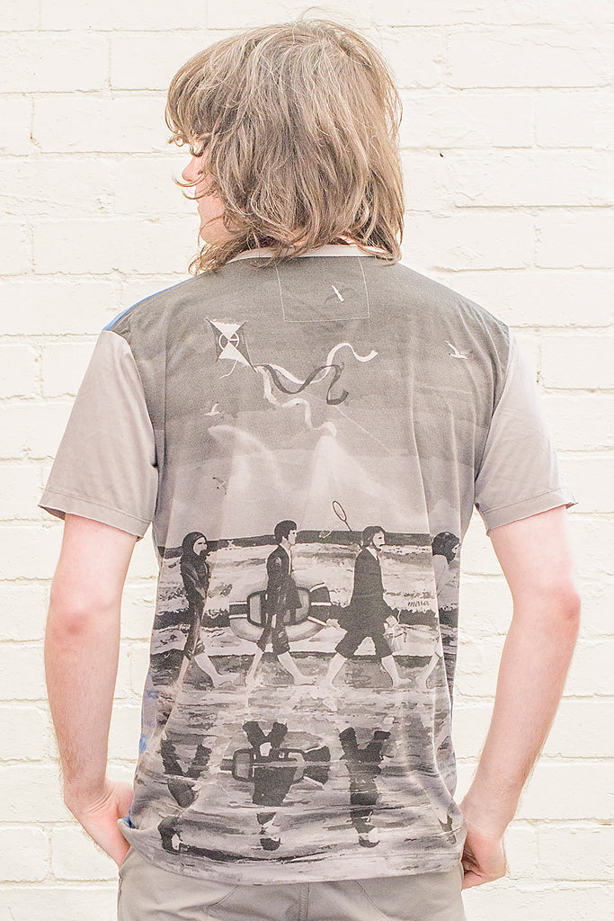 model wearing a grey T-shirt featuring a painterly design of the beatles walking along a beach with beach toys from the back