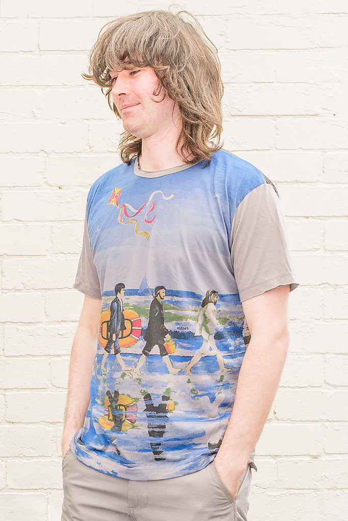 model wearing a grey T-shirt featuring a painterly design of the beatles walking along a beach with beach toys from the front