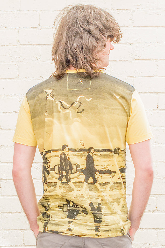 model wearing a yellow T-shirt featuring a painterly design of the beatles walking along a beach with beach toys back