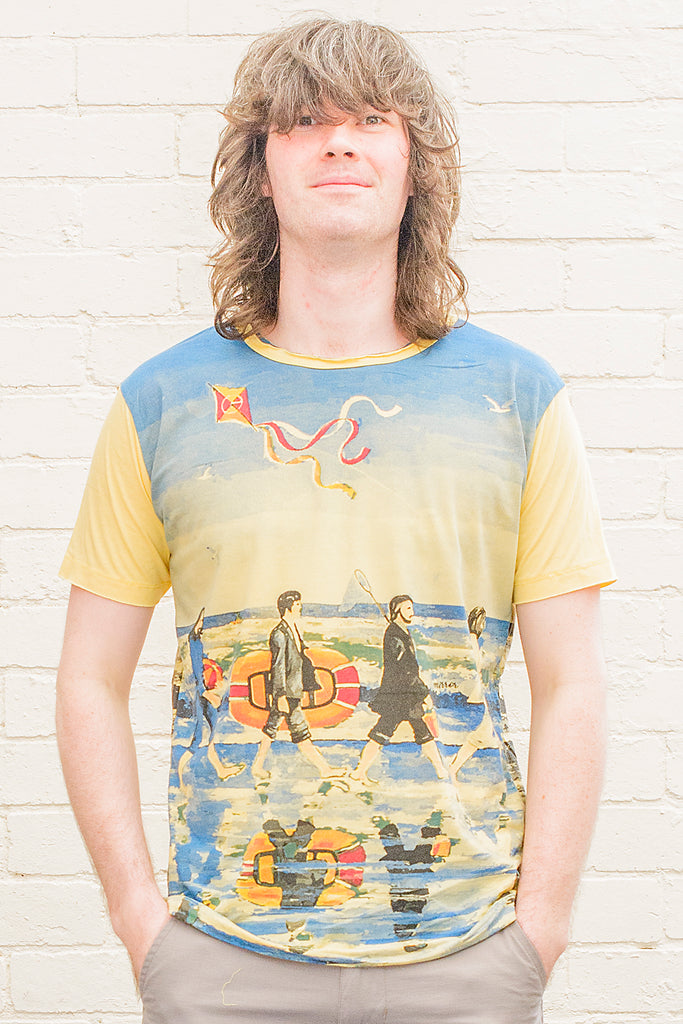 model wearing a yellow T-shirt featuring a painterly design of the beatles walking along a beach with beach toys