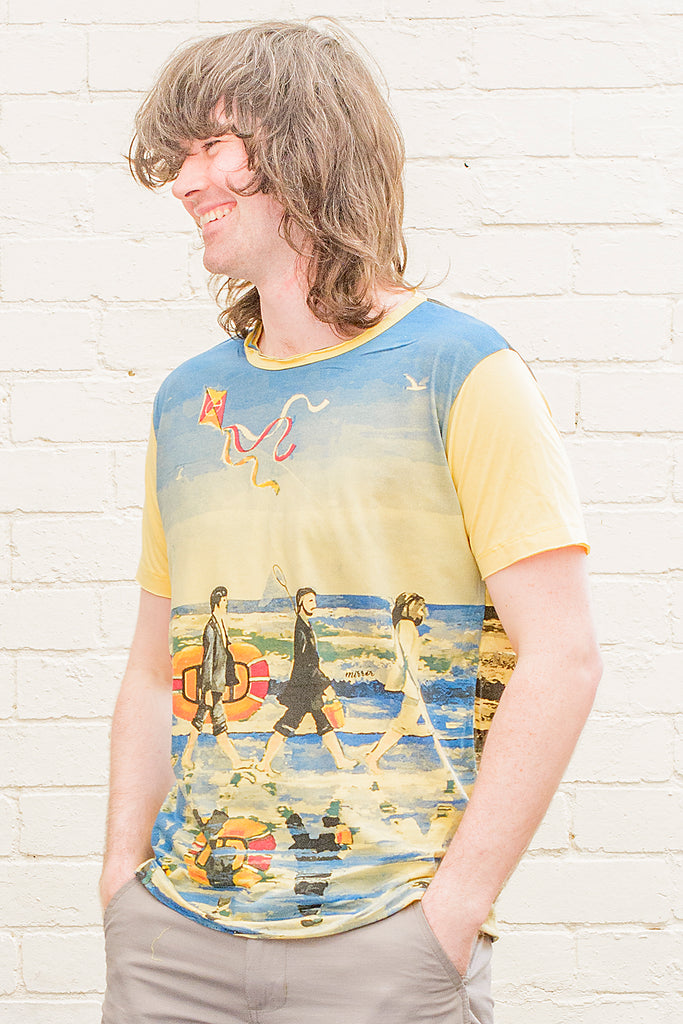 model wearing a yellow T-shirt featuring a painterly design of the beatles walking along a beach with beach toys