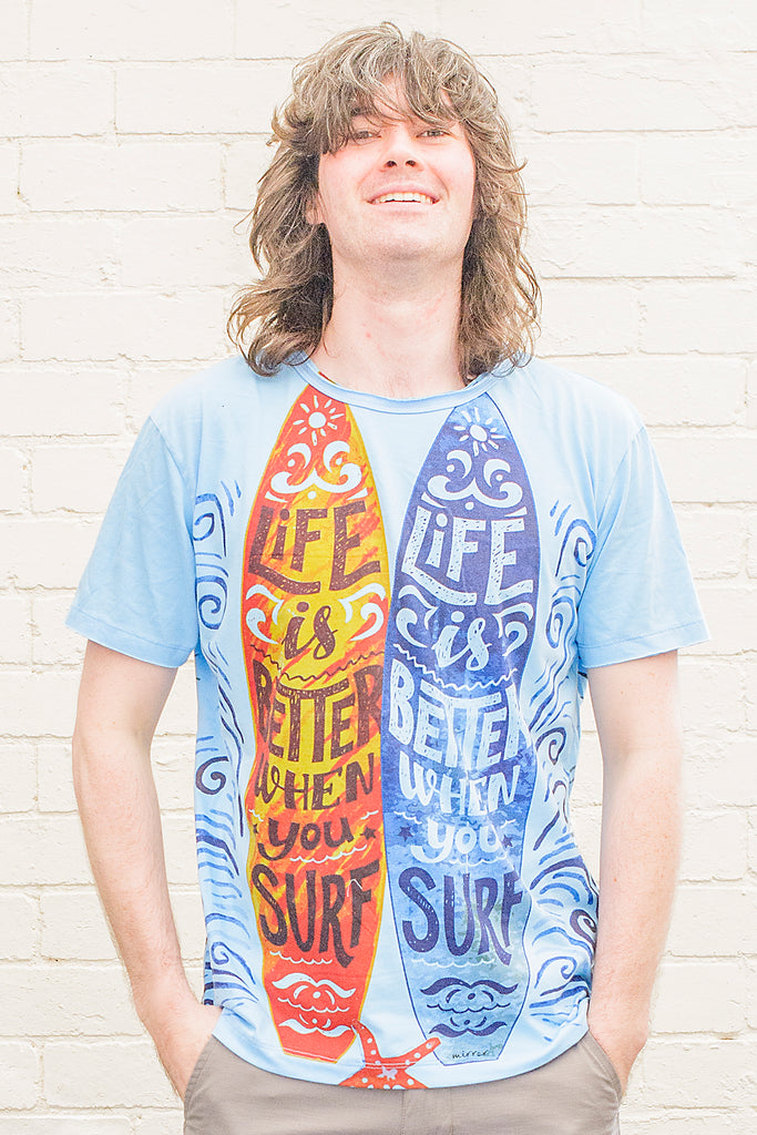 model wearing a bright blue t-shirt with a graphical print of two surfboards sticking out of the sand at the beach with life is better when you surf lettering on them from the front