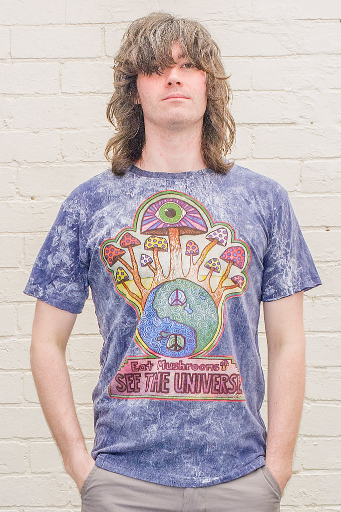 model wearing a blue stonewash t-shirt printed with a colourful magic mushroom graphic and eat mushrooms see the universe lettering from the front